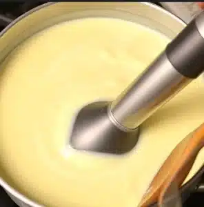 Creamy mashed potato soup simmering in a pot on the stove, with an immersion blender blending it smooth. Steam rises from the hot soup, and a wooden spoon rests nearby.
